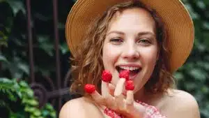 Close up young beautiful happy woman 20s wear pink dress hat put girl put raspberries on fingers eat stand outdoor near forged gate on green ivy background People urban summer time lifestyle concept.