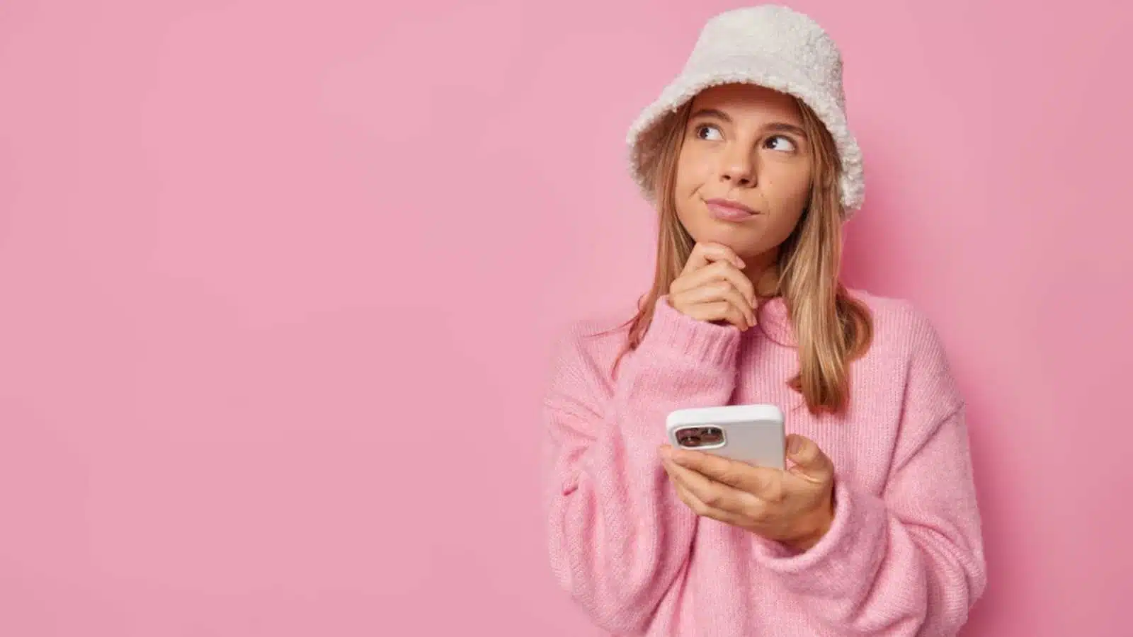 Doubtful young lovely woman holds chin thinks about mobile online offer wears hat and casual jumper looks away contemplates about something isolated over pink background with copy space on left