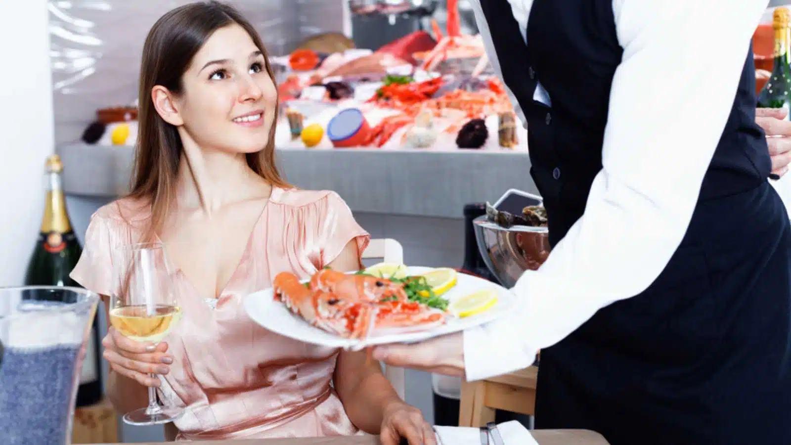 Polite waiter serving ordered seafood dishes to attractive cheerful smiling girl at fish restaurant