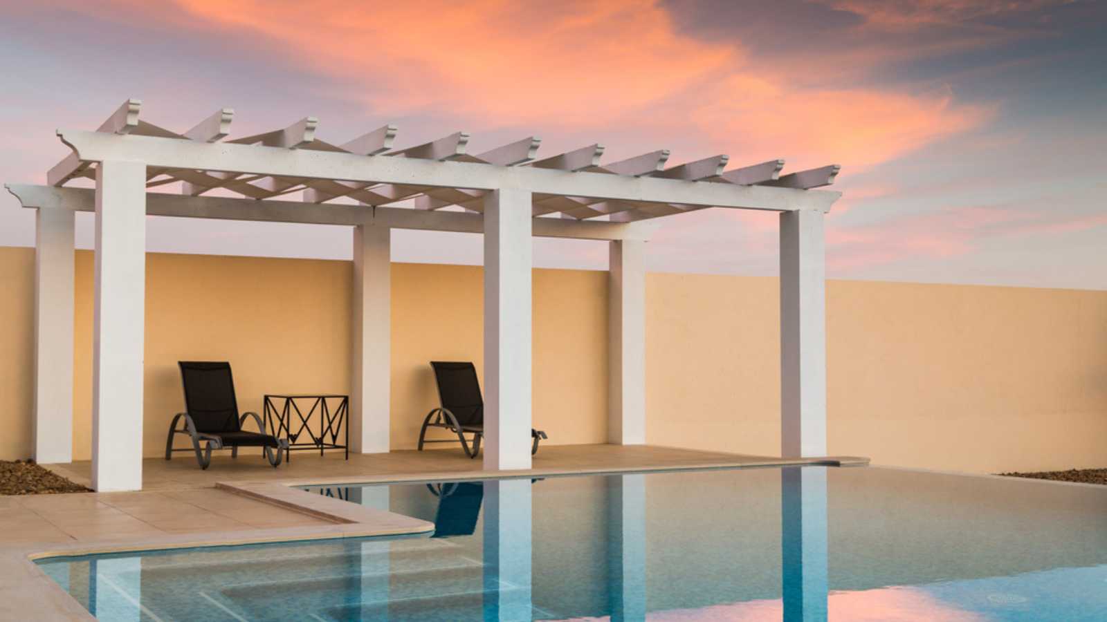 White poolside pergola, gazebo providing shade on a terrace patio area next to an infinity swimming pool at dusk as the sunset turns the sky pink orange.