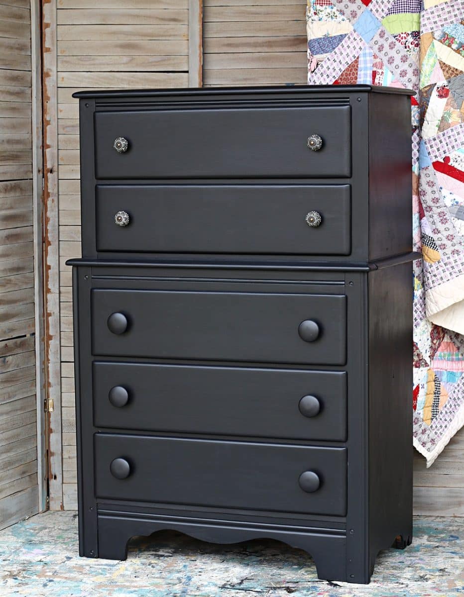 A black-painted vintage dresser with two smaller drawers on top and three larger drawers below, featuring round knobs, stands in front of wooden shutters and a colorful patchwork quilt.