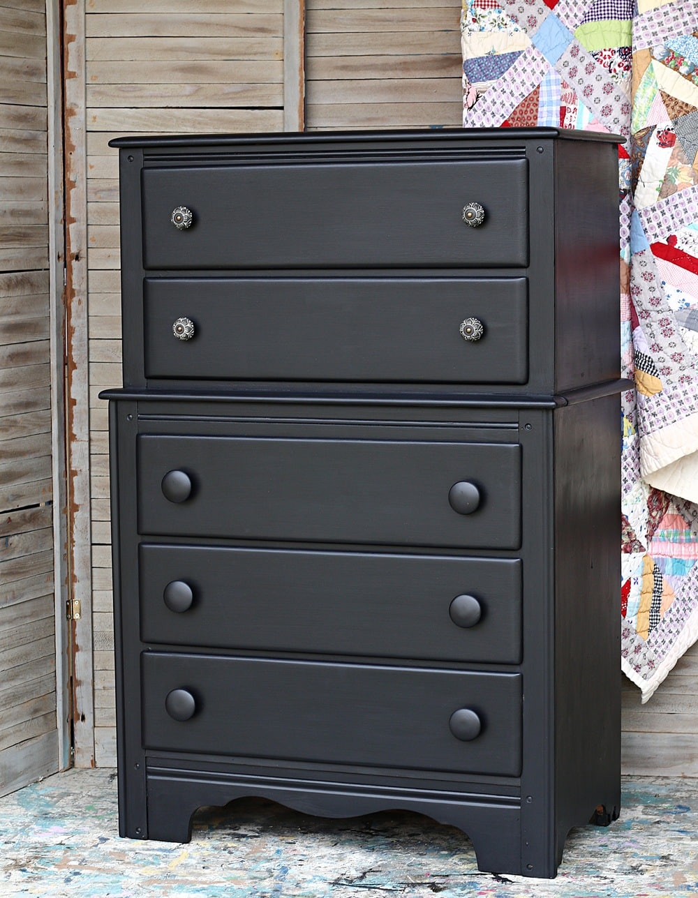 A black-painted vintage dresser with two smaller drawers on top and three larger drawers below, featuring round knobs, stands in front of wooden shutters and a colorful patchwork quilt.