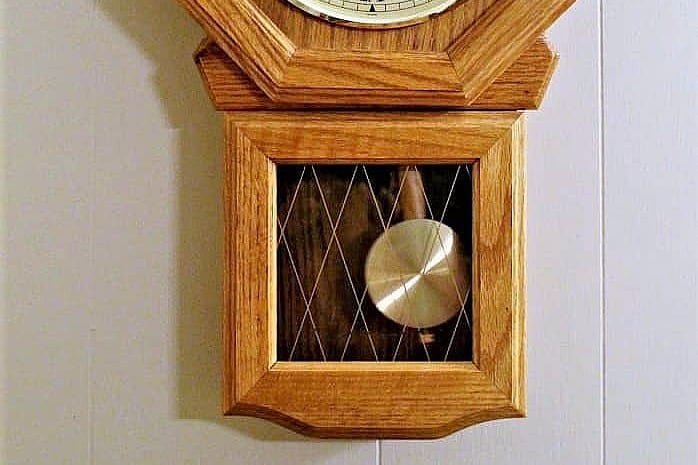A close-up of the lower half of a wooden wall clock with a glass front panel featuring a gold diamond pattern, showing the metal pendulum inside.