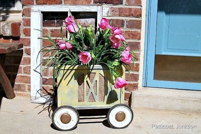A yellow vintage cart with white wheels holds pink tulips and green leaves, placed in front of a brick wall and a blue door. An empty white window frame leans against the wall behind the cart.